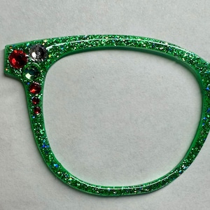 Puede incluir: Montura de gafas verde brillante con gemas decorativas rojas, transparentes y verdes en el lado izquierdo. La montura es de color verde brillante y festivo, perfecta para un disfraz o una fiesta.