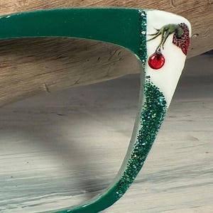 Puede incluir: Montura de gafas verde con una sección blanca con un diseño temático de Grinch y un adorno rojo. La montura tiene detalles de purpurina verde a lo largo del borde. Las gafas están sobre un fondo de madera clara.