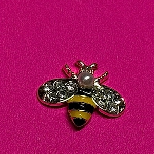 Puede incluir: Broche de abeja dorada con rayas negras y amarillas en el cuerpo y detalles de cristal transparente en las alas. Una pequeña perla se encuentra en la parte superior de la cabeza de la abeja. El broche está sobre un fondo rosa brillante.