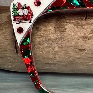 May include: A festive eyeglass frame with a white section featuring a Christmas scene with a red truck, tree, and gnomes. The frame is red with embedded red and green glitter, and has three red jewel accents.