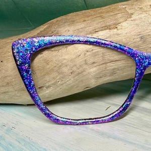 Può includere: Una montatura di occhiali a forma di occhi di gatto scintillante. La montatura è un vibrante mix di blu, viola e glitter iridescenti, che crea un effetto abbagliante. La montatura è posta su uno sfondo di legno naturale.