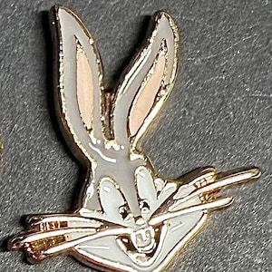 May include: A Bugs Bunny enamel pin. The pin features the iconic cartoon rabbit with a gray body, pink inner ears, and a carrot in his mouth. The pin has a gold-colored outline and is set against a gray background.