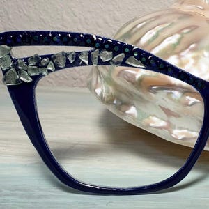 May include: Close-up of a pair of navy blue eyeglasses with a cat-eye frame. The upper frame is embellished with clear, irregularly shaped stones and small, dark green accents. The glasses are set against a blurred, pearlescent background.