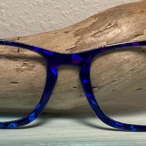 Peut inclure: Lunettes bleues avec un motif tacheté. Les montures ont une forme rectangulaire et sont posées sur un fond en bois. Les lunettes sont d'un bleu vif avec des touches de violet.