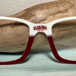 May include: White and crimson eyeglasses with the word "Alabama" and the letter "A" printed on the frames. The frames have a thick, bold design with a white top and crimson bottom. The eyeglasses are set against a wooden background.