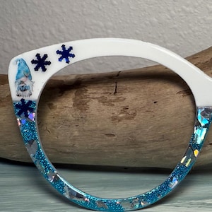 May include: White eyeglass frame with a blue glitter and snowflake design. The frame features a gnome illustration on the upper left side. The lower portion of the frame is filled with blue glitter and iridescent pieces.