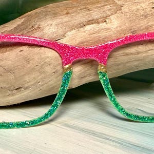 May include: Pink and green glitter eyeglasses with a watermelon design. The top of the frames are pink with black accents, transitioning to green at the bottom. The glasses are resting on a piece of driftwood.