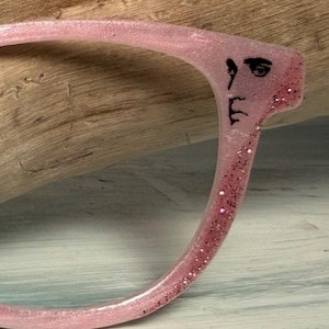 May include: A pink eyeglass frame with glitter and a black face design. The frame is made of a translucent material and has a curved shape. The face design is on the temple of the frame.