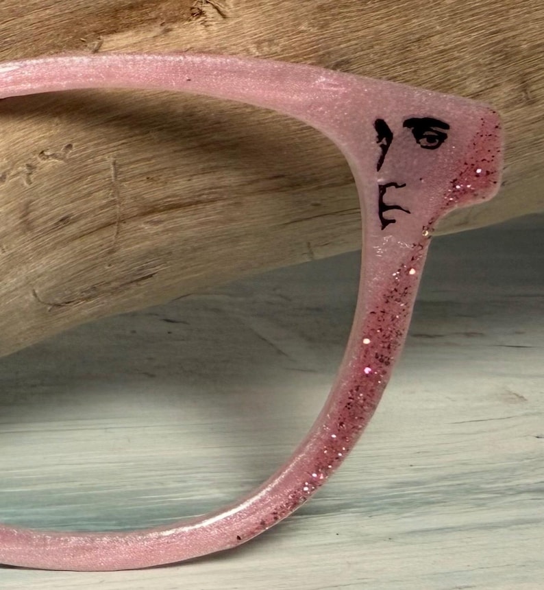 May include: A pink eyeglass frame with glitter and a black face design. The frame is made of a translucent material and has a curved shape. The face design is on the temple of the frame.
