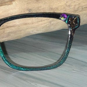 May include: A single eyeglass frame with a glittery teal and black finish. The frame features a decorative purple gem and a gold-colored embellishment on the corner.