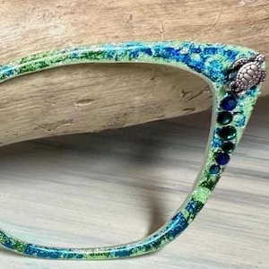 May include: A decorative eyeglass frame with a sea-themed design. The frame features a blend of green and blue glitter, with a silver turtle accent on the corner and blue gemstones. The frame is set against a wooden background.