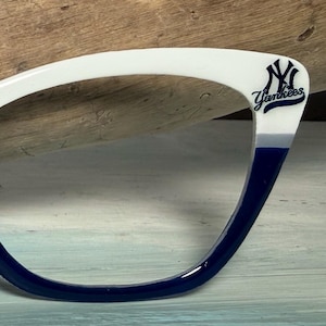 Puede incluir: Montura de gafas blanca y azul marino con el logotipo de los Yankees de Nueva York. La montura tiene una sección superior blanca y hace la transición a azul marino en la parte inferior. El logotipo de los Yankees es azul marino.