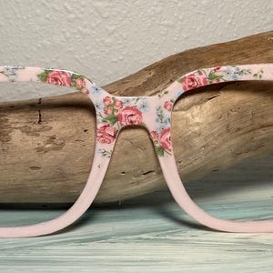 Peut inclure: Monture de lunettes rose pâle ornée d'un motif floral de roses roses et bleues. Les montures sont posées sur un fond en bois naturel, mettant en valeur le design délicat et la palette de couleurs.