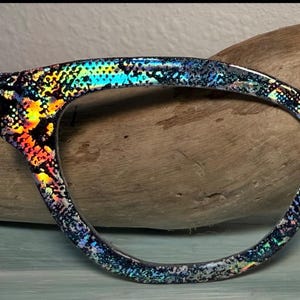 May include: Close-up of a pair of eyeglasses frames. The black frames feature a vibrant, iridescent pattern with hues of yellow, orange, blue, and green. The speckled design gives a textured appearance, creating a unique and striking look.