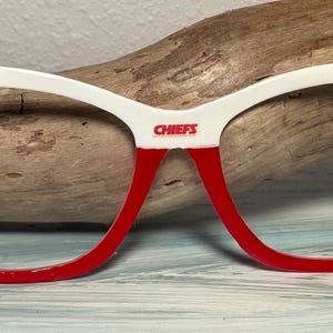 May include: White and red cat-eye glasses with the word "CHIEFS" printed on the bridge. The glasses have red frames and white upper rims. Small images of football players are on the upper corners.