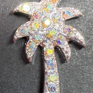Op de afbeelding: Zilverkleurige palmboom broche met iriserende, kleurrijke strass-steentjes. De broche heeft een gedetailleerd ontwerp van een palmboom met een getextureerd oppervlak en een sluiting met speld. Een accessoire met tropisch thema.