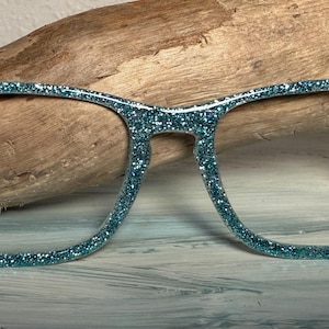Può includere: Occhiali glitterati color teal con montatura rettangolare. Le montature sono ricoperte di glitter scintillanti e presentano una piccola gemma blu su ogni angolo. Gli occhiali sono appoggiati su uno sfondo di legno.