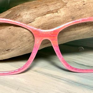 May include: Pink cat-eye style eyeglasses with a glittery finish. The frames feature black palm tree and bird silhouettes on the upper corners. The glasses are set against a wooden background.