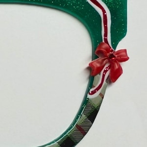 Könnte beinhalten: Ein grüner, glitzernder, dekorativer Rahmen mit einer roten Schleife und einem weiß-roten Streifenakzent. Der Rahmen hat unten ein Karomuster. Das Design ist festlich und könnte für Weihnachtsdekorationen verwendet werden.