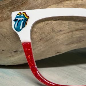 May include: White-framed spectacles with a red glitter accent on the lower frame. The Rolling Stones tongue logo is on the upper corner. The glasses are set against a wood grain background.