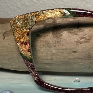 Op de afbeelding: Een decoratief brilmontuur met een glitterend harsontwerp. Het montuur heeft een kleurengradatie van goud, groen en bordeauxrood. Het ligt op een stuk drijfhout.
