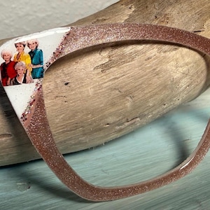 May include: A rose gold glitter cat-eye eyeglass frame. The left side features a white section with a photo of the Golden Girls cast. The frame rests on a piece of driftwood.