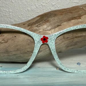 May include: Cat-eye style eyeglasses with a glittery, light blue frame. The glasses feature red ladybug and flower accents. The lenses are clear. The glasses are set against a wooden background.