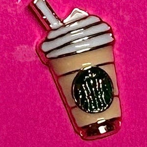 May include: A gold-colored enamel pin in the shape of a coffee drink with whipped cream. The pin features a green logo and is set against a bright pink background.