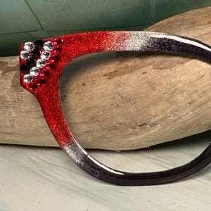Puede incluir: Una montura de gafas decorativa con un acabado de purpurina roja que se desvanece a plata y negro. La montura está adornada con pedrería transparente y negra. Un accesorio único.