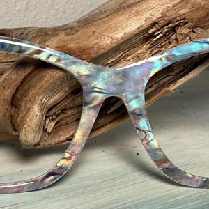 May include: Cat-eye style eyeglasses with a pearlescent, iridescent finish. The frames feature a white accent on the upper corners, embellished with a small, dark-colored turtle design. The glasses are set against a natural wood background.