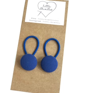 May include: Two navy blue fabric-covered button hair ties with blue elastic loops. The hair ties are attached to a brown card with a white label that has the text "little thimbles" and Etsy and Instagram information.