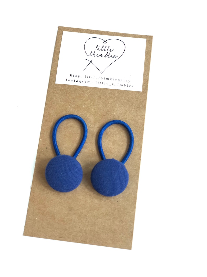 May include: Two navy blue fabric-covered button hair ties with blue elastic loops. The hair ties are attached to a brown card with a white label that has the text "little thimbles" and Etsy and Instagram information.