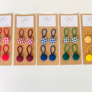 May include: A collection of fabric-covered button hair ties in various colors and patterns. Each set includes two hair ties, with options like red, burgundy, blue, green, and yellow. Some buttons feature a gingham pattern.