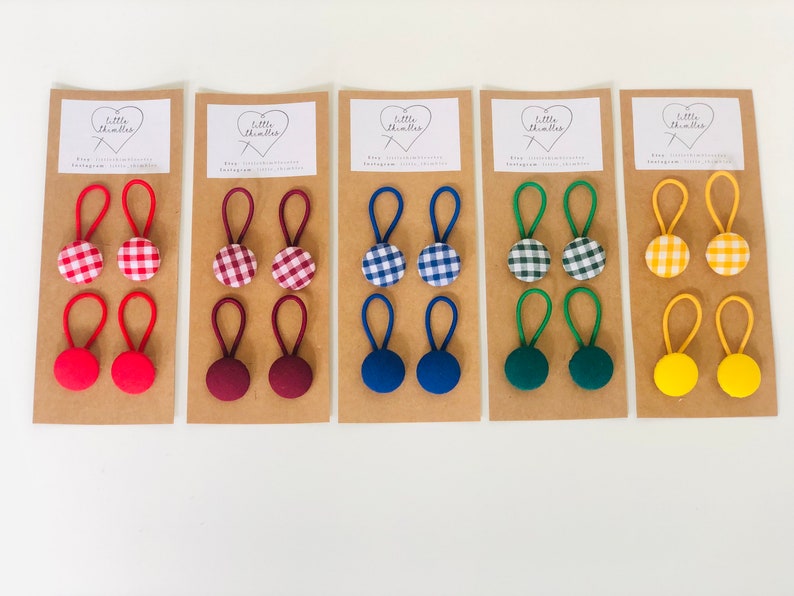 May include: A collection of fabric-covered button hair ties in various colors and patterns. Each set includes two hair ties, with options like red, burgundy, blue, green, and yellow. Some buttons feature a gingham pattern.