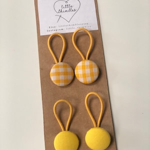 May include: Four yellow fabric buttons with elastic loops attached to a brown card. Two buttons have a yellow and white gingham pattern, and the other two are solid yellow. The card has the text "little thimbles" and Etsy and Instagram information.