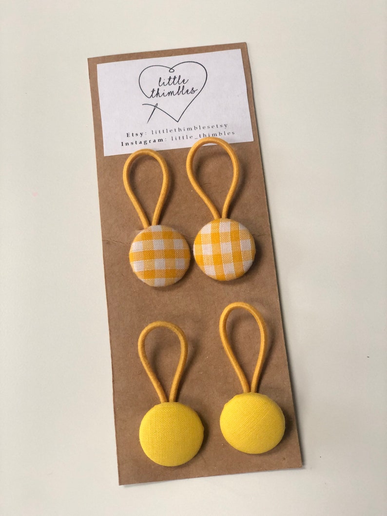May include: Four yellow fabric buttons with elastic loops attached to a brown card. Two buttons have a yellow and white gingham pattern, and the other two are solid yellow. The card has the text "little thimbles" and Etsy and Instagram information.
