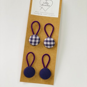 May include: Four hair ties on a brown card. Two ties have purple gingham fabric buttons, and two have solid navy blue fabric buttons. The ties have purple elastic loops. The card has a white label with a heart and the text "little thimbles".