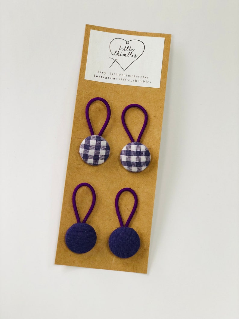 May include: Four hair ties on a brown card. Two ties have purple gingham fabric buttons, and two have solid navy blue fabric buttons. The ties have purple elastic loops. The card has a white label with a heart and the text "little thimbles".