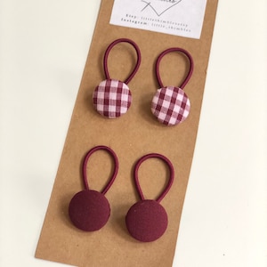 May include: Four burgundy hair ties with elastic loops and fabric-covered buttons. Two ties have a gingham pattern in burgundy and white, while the other two are solid burgundy. The ties are displayed on a brown card with the text "little thimbles".