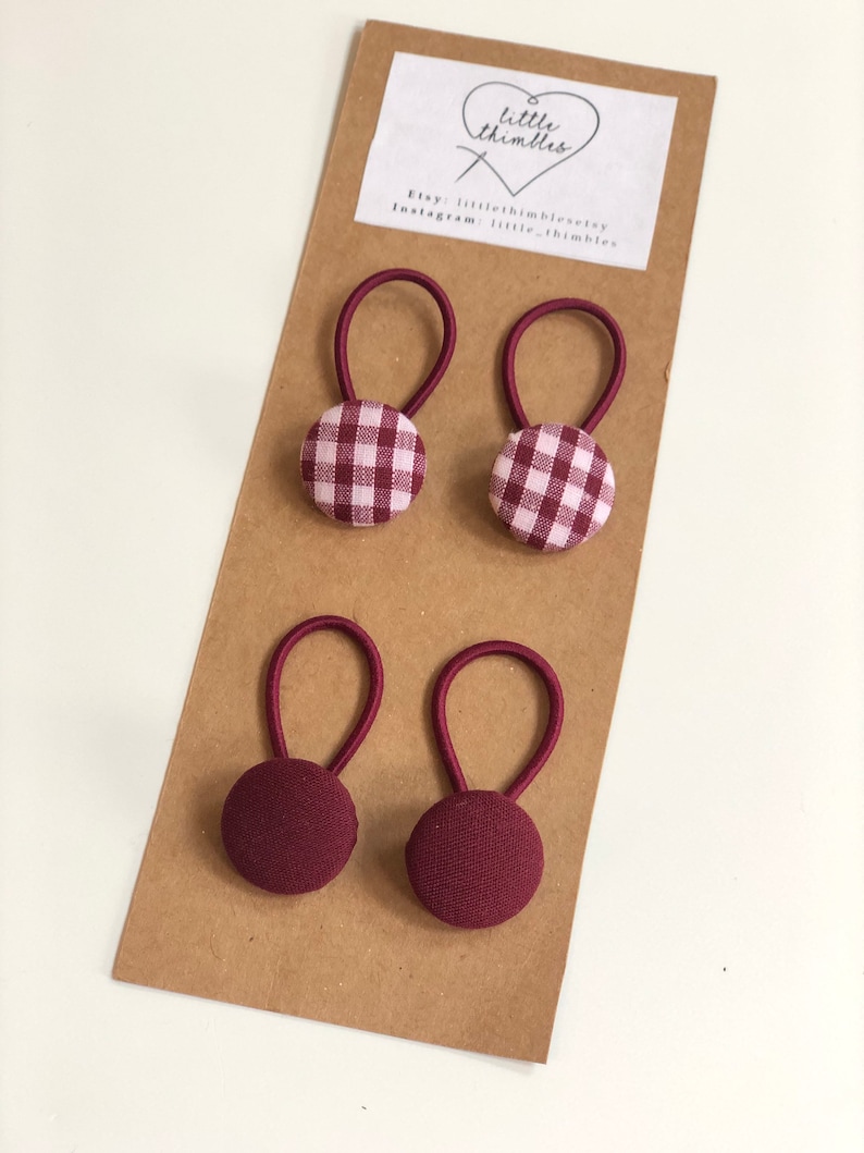 May include: Four burgundy hair ties with elastic loops and fabric-covered buttons. Two ties have a gingham pattern in burgundy and white, while the other two are solid burgundy. The ties are displayed on a brown card with the text "little thimbles".