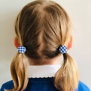 May include: A child's blonde hair styled in two pigtails, secured with blue and white gingham fabric-covered hair ties. The child is wearing a blue sweater with a white collared shirt underneath.