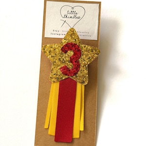 May include: A gold glitter star badge featuring a red glitter number 3 in the centre, attached to a brown card with the text little thimbles. Below the star are three vertical ribbons in alternating yellow and red colours hanging downwards.
