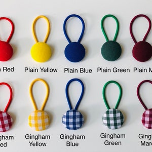May include: A collection of ten fabric-covered button hair ties in various colors and patterns. The top row features solid colors: red, yellow, blue, green, and maroon. The bottom row displays gingham patterns in red, yellow, blue, green, and maroon. Each tie has a loop.