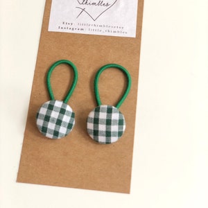 May include: Two green and white gingham button hair ties with green elastic loops are displayed on a brown card. The card has a white label with the text "little thimbles" and Etsy and Instagram account information.