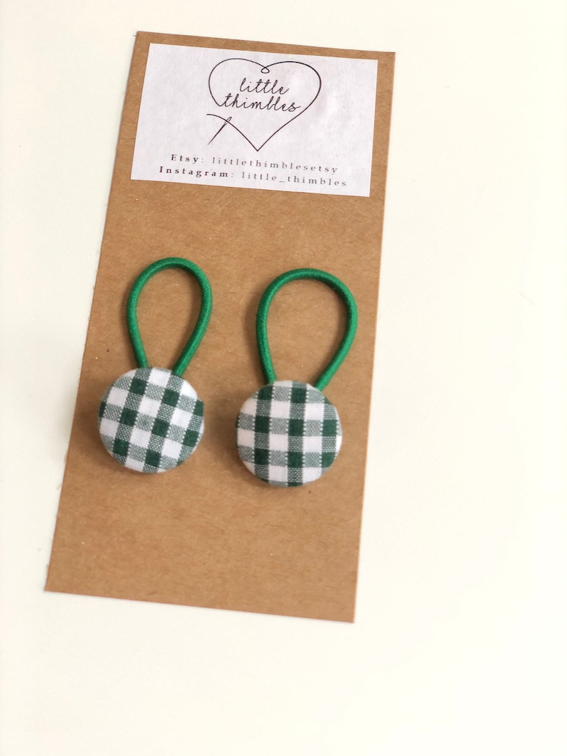 May include: Two green and white gingham button hair ties with green elastic loops are displayed on a brown card. The card has a white label with the text "little thimbles" and Etsy and Instagram account information.