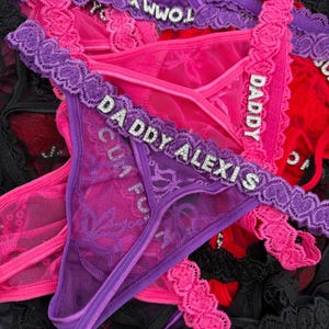 May include: A collection of colorful thong underwear in shades of pink, purple, and black. The lingerie features lace detailing and rhinestone lettering spelling out phrases like "Daddy Alexis" and "Tommy".