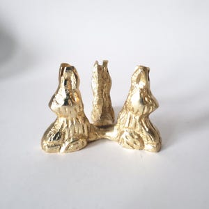 May include: A gold-colored, abstract sculpture with three vertical elements connected at the base. The textured surface reflects light, creating highlights and shadows. The design is organic and artistic.