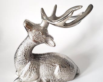 May include: A silver-toned metal figurine of a deer in a resting position. The deer has textured fur detailing across its body and head, with prominent antlers extending backward. The surface of the metal is polished and reflective.