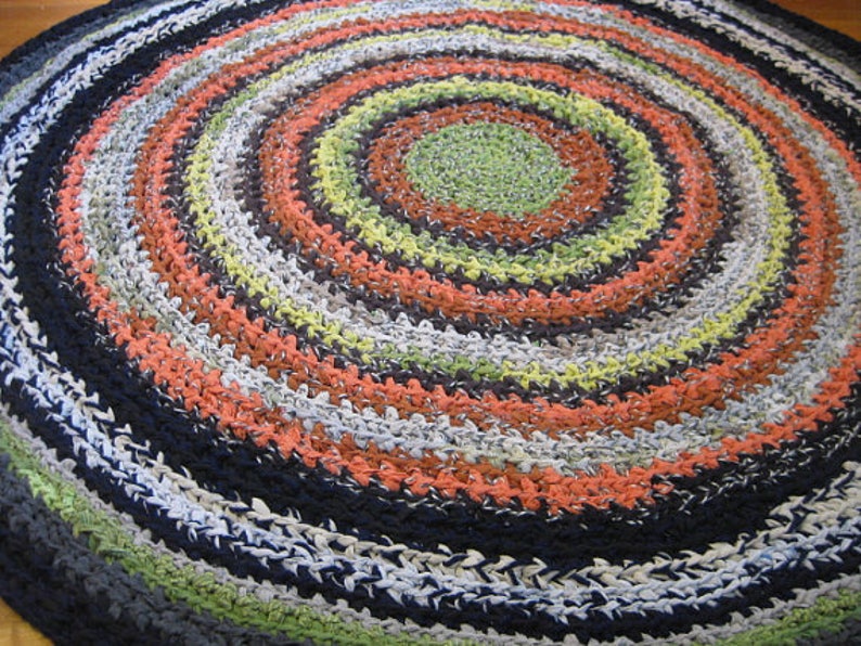 Op de afbeelding: Een handgemaakt, cirkelvormig tapijt met een spiraalvormig ontwerp. Het tapijt heeft concentrische ringen in verschillende kleuren, waaronder groen, oranje, zwart, wit en grijs. Het tapijt is gemaakt van een textuurrijk materiaal, waarschijnlijk stof of garen.