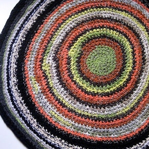 Op de afbeelding: Een ronde, handgemaakte vloerkleed met een spiraalvormig ontwerp. Het vloerkleed heeft concentrische cirkels in verschillende kleuren, waaronder zwart, grijs, wit, oranje, groen en bruin. Het vloerkleed is gemaakt van garen en heeft een getextureerd oppervlak.